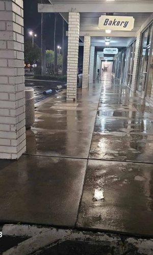 Pressure washing sidewalk in front of bakery and retail stores at night.